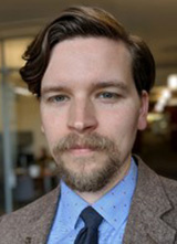 Man with brown hair wearing a brown jacket, blue dotted shirt, and black tie 